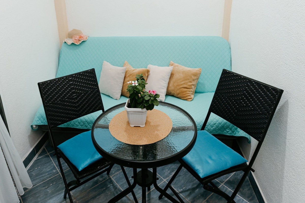 A small, enclosed outdoor seating area with a round glass table, two chairs, and a light blue cushioned sofa with multiple pillows.