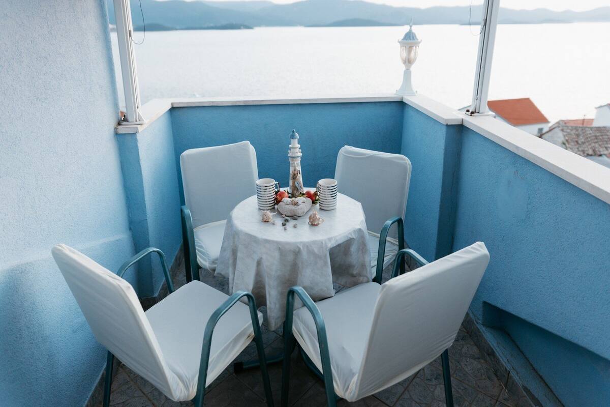 A small round table with a white tablecloth, surrounded by four white chairs, set against a blue wall with a view of the sea and distant mountains.