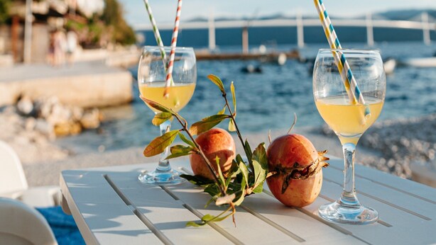 Two glasses of yellow beverage with striped straws, a pomegranate, and a branch on a white table by a waterfront. - Apartments Dragobratović (Slivno)