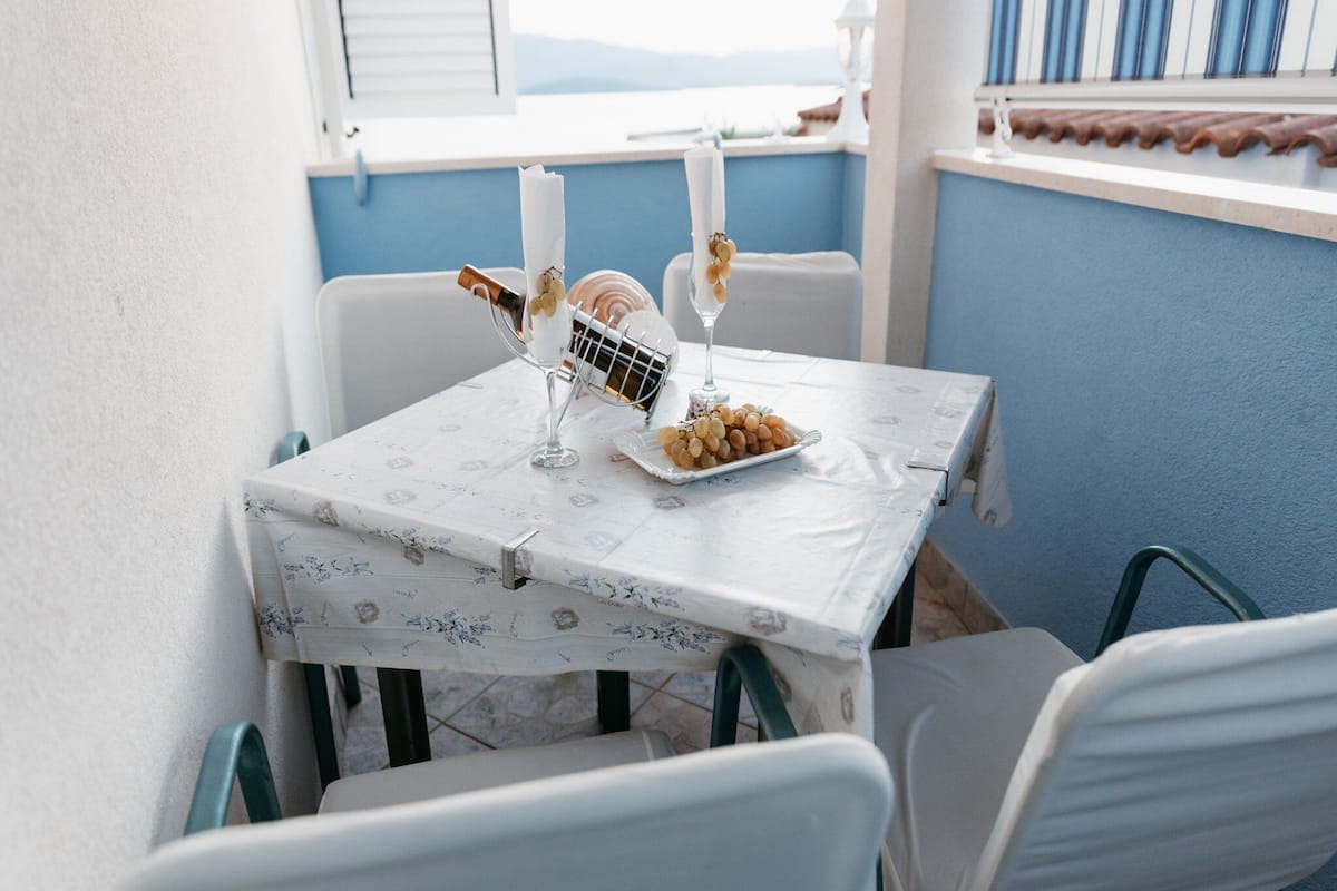 A small outdoor dining area with a table set for two, featuring a white tablecloth, a plate of grapes, and a candle holder.