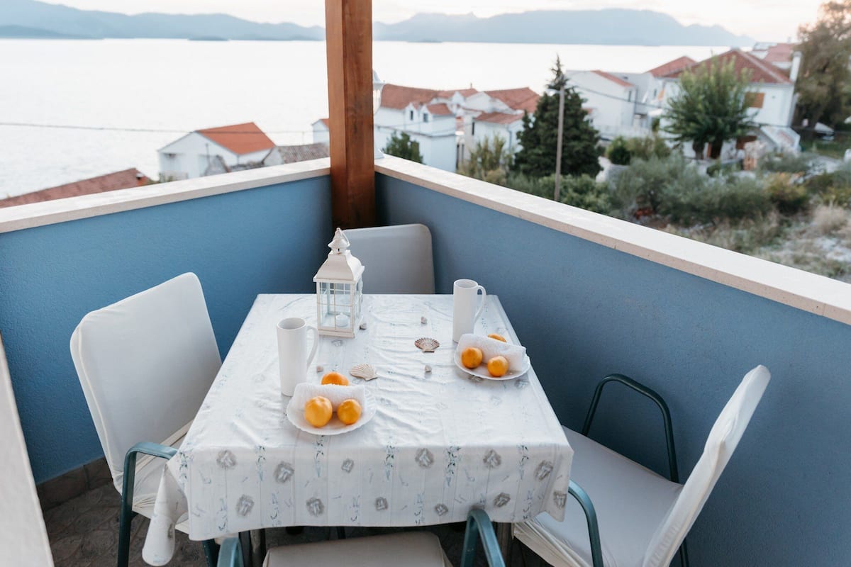 A table set with a white tablecloth, two plates with food, a white lantern, and a cup on a balcony overlooking a body of water and buildings.
