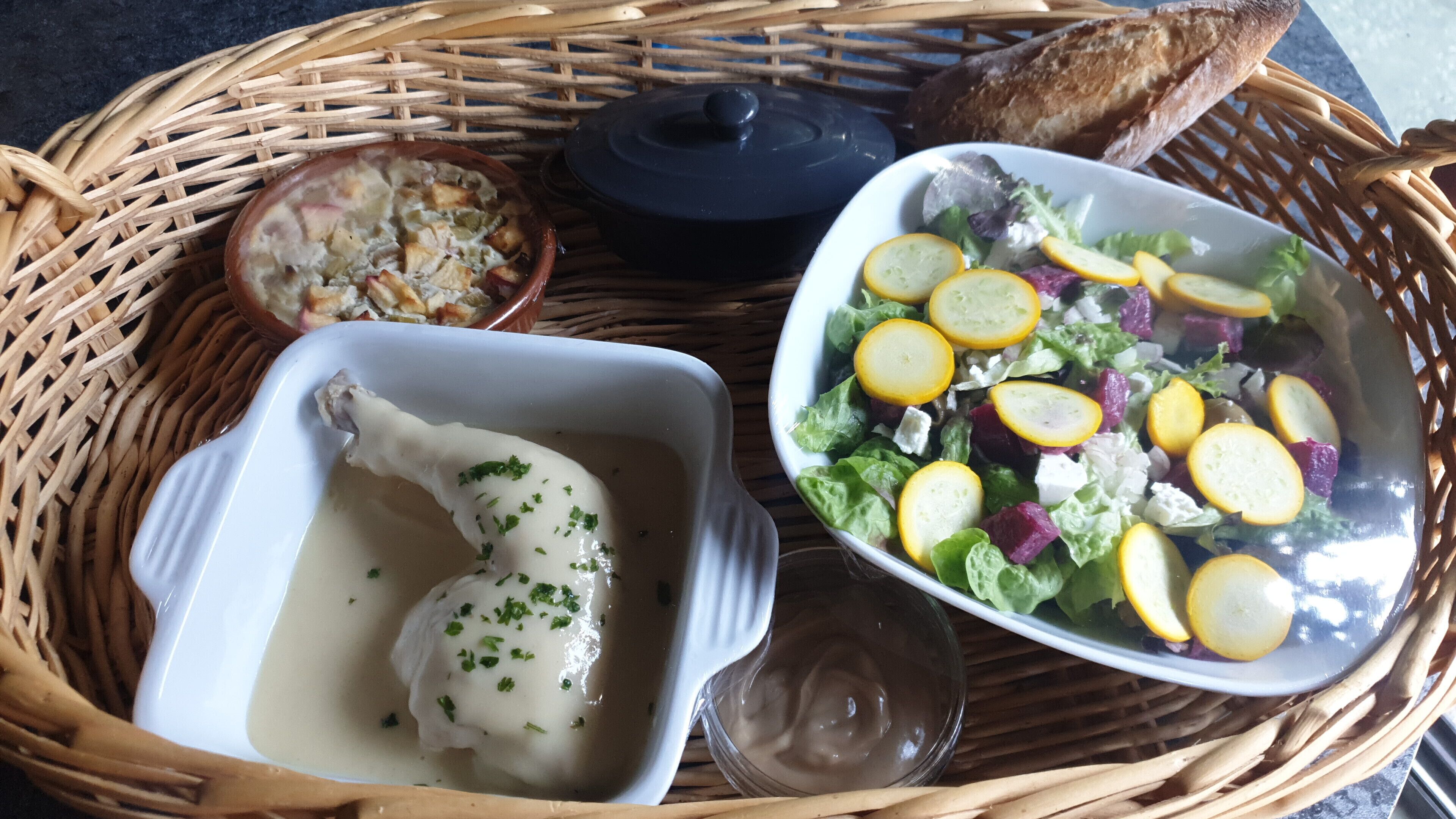 Un cesto di vimini con una ciotola bianca contenente un osso, un'insalata con fette di limone e una pentola scura.