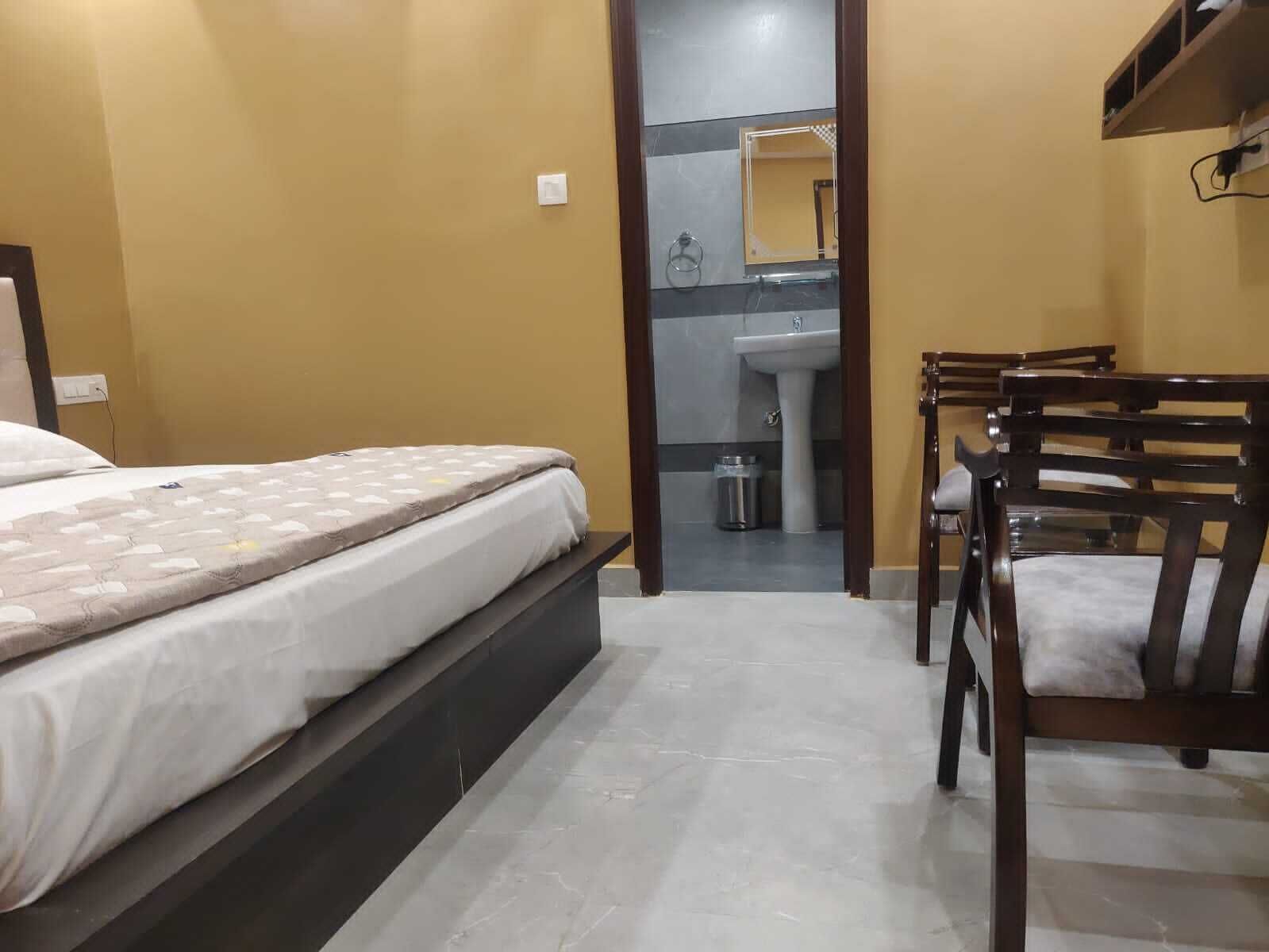 Un dormitorio con cama, una silla, una mesita y un baño visible a través de una puerta abierta.
