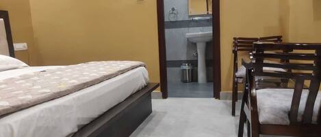 Un dormitorio con cama, una silla, una mesita y un baño visible a través de una puerta abierta.