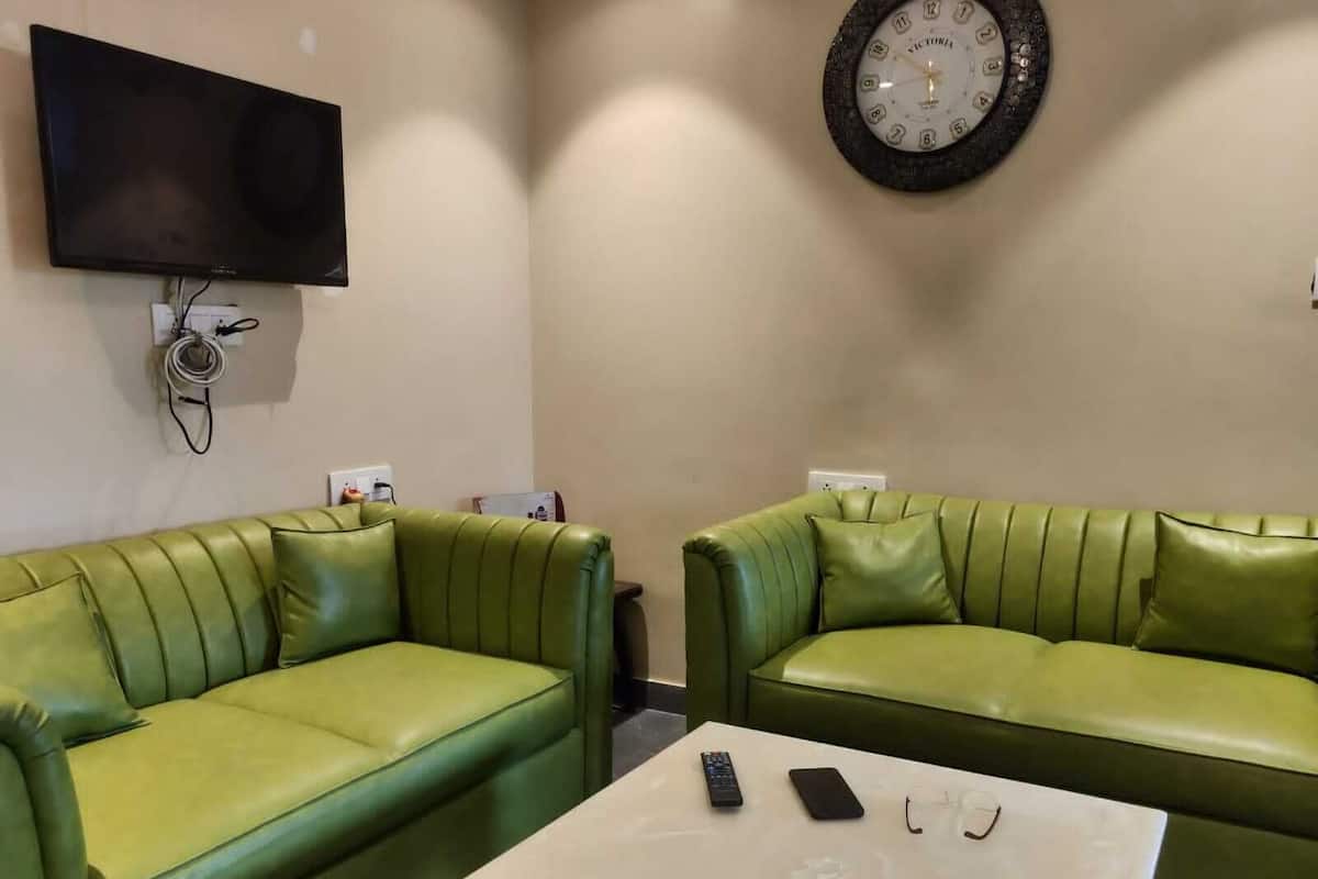 A room with two green leather sofas, a flat-screen TV mounted on the wall, a round wall clock, and a table with a remote control, smartphone, and glasses.