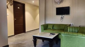 A living room with a green sofa, a white coffee table, a flat-screen TV mounted on the wall, and a ceiling fan. - The Republic Hotel (Ambala)
