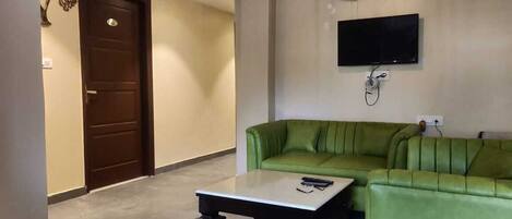 A living room with a green sofa, a white coffee table, a flat-screen TV mounted on the wall, and a ceiling fan.