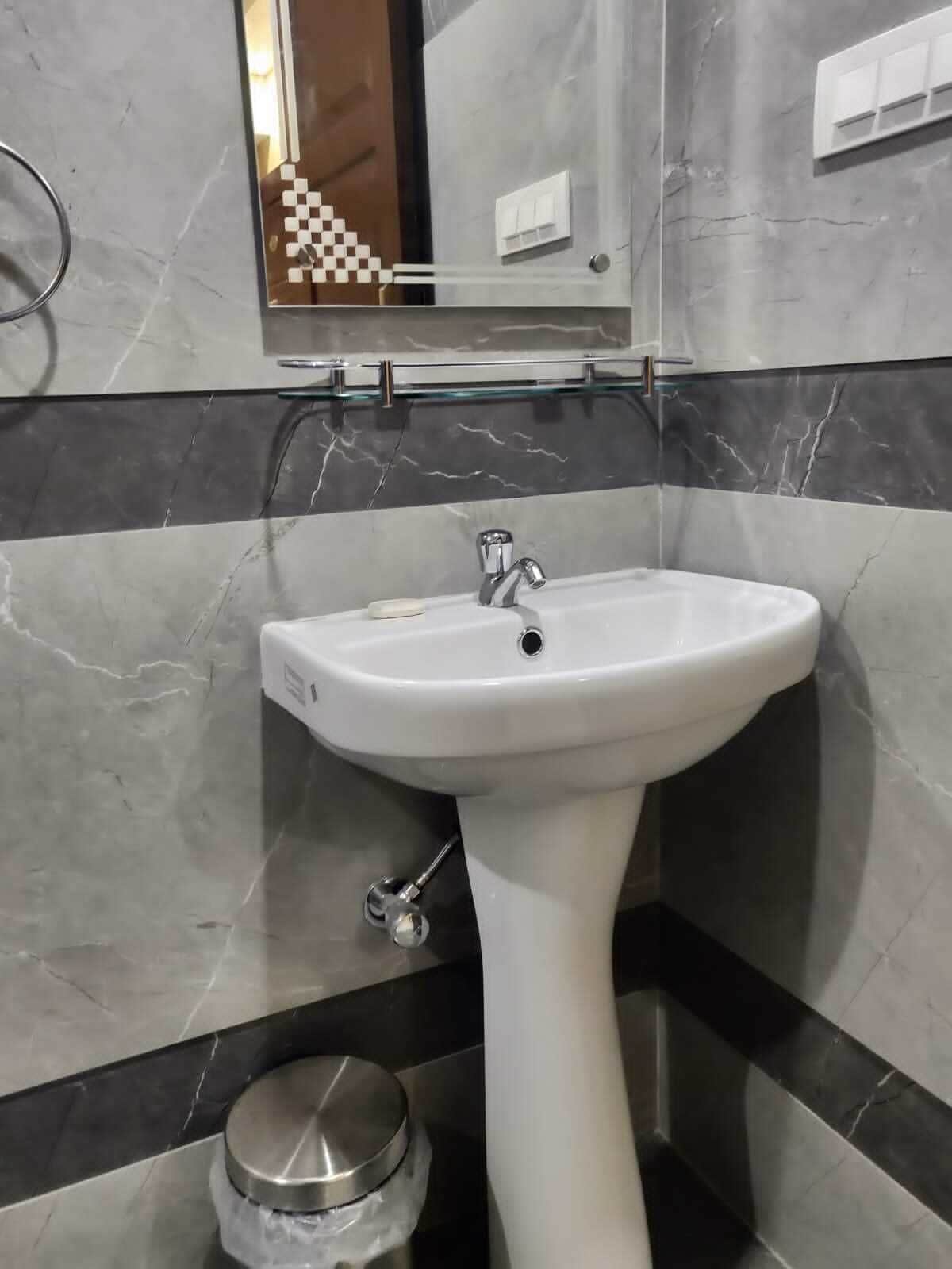 Ein Badezimmer mit einem weißen Waschtisch, einer Glasablage über dem Spiegel und einer Wand mit Marmoroptik.