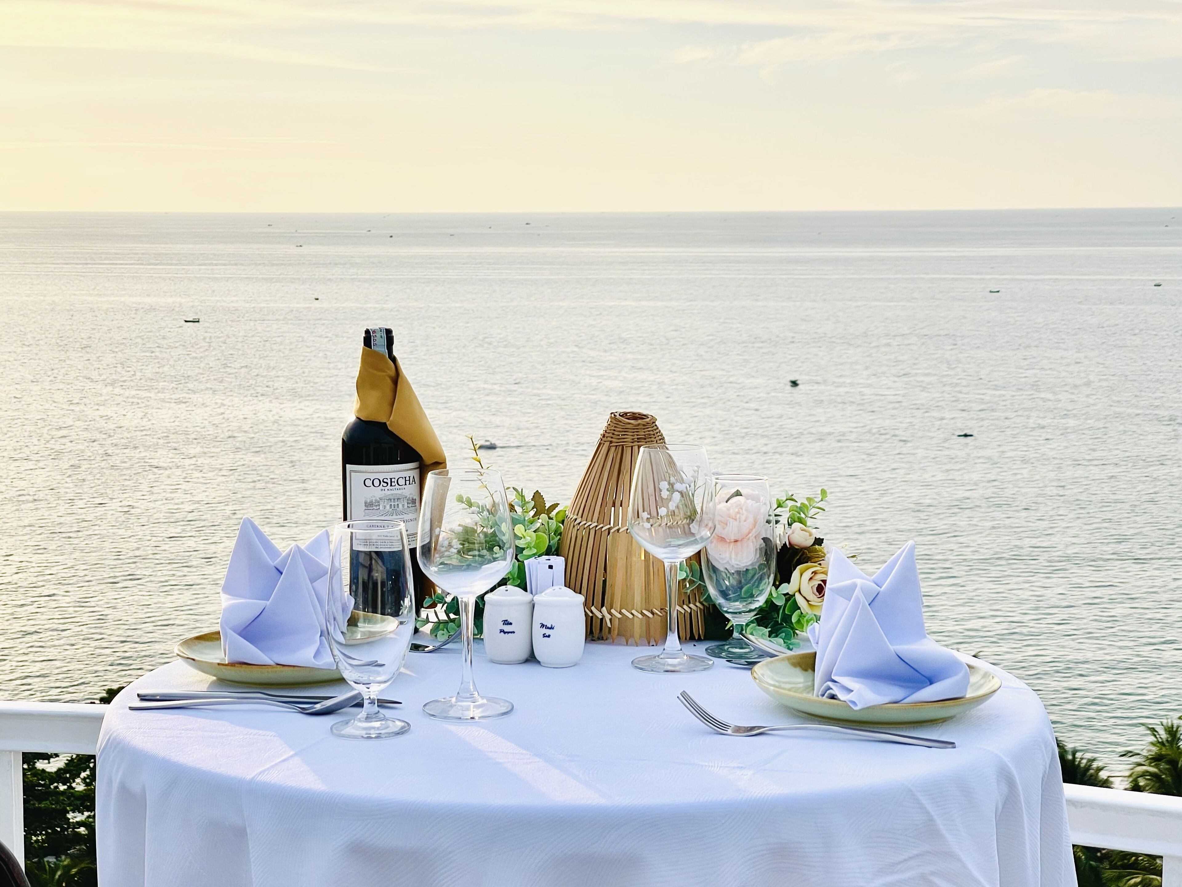 A table set for two with a bottle of wine, glasses, and a decorative centerpiece overlooking the sea.