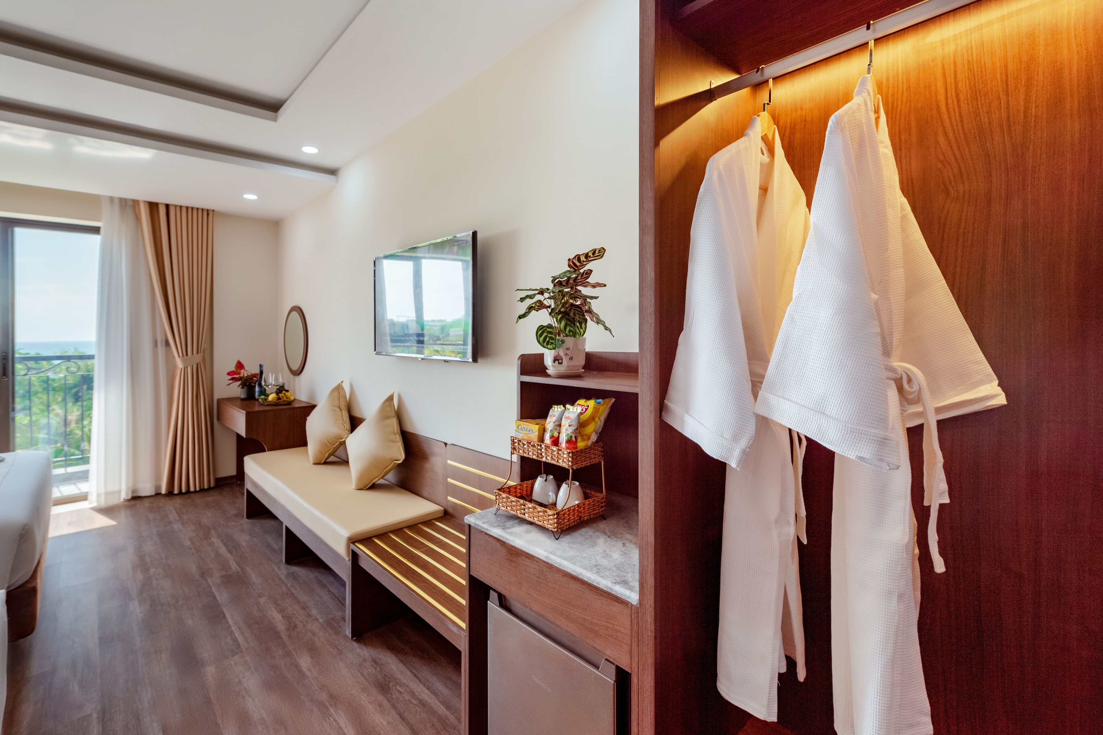 A modern hotel room with a wooden floor, a sofa, a small desk, a mirror, a TV, and a wardrobe with hanging clothes.
