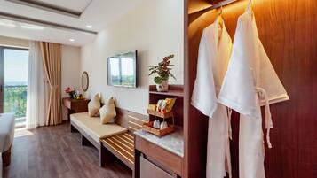 A modern hotel room with a wooden floor, a sofa, a small desk, a mirror, a TV, and a wardrobe with hanging clothes.