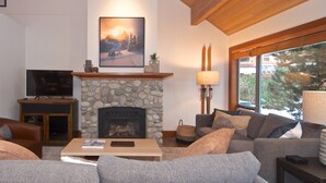 A cozy living room with a stone fireplace, a mounted TV, and a large window with a view of trees.