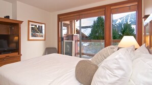 A hotel room with a large bed, a TV, a framed picture, and a view of mountains through the windows.
