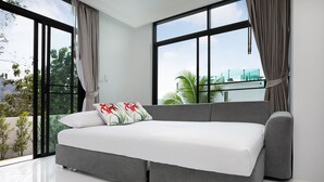 A modern bedroom with a grey sofa bed, large windows, and a view of greenery and a building. - King Size Sea View Villa + Pool + Rooftop Skydeck  (Kathu)