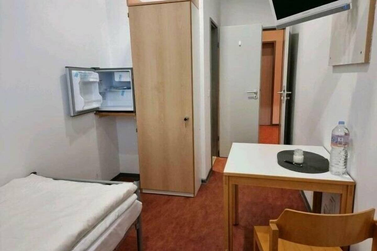 A small, compact room with a single bed, a desk with a chair, and a water bottle on the desk.