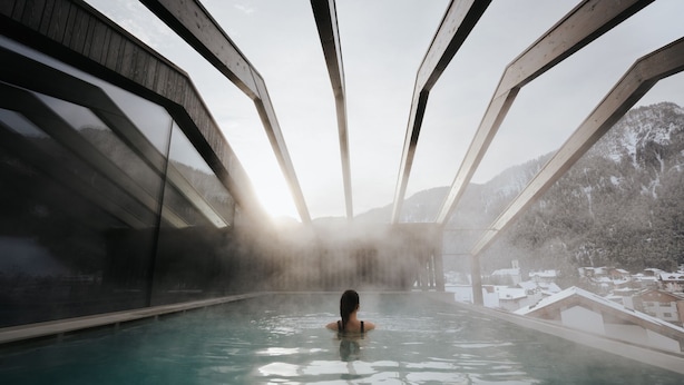 Una persona en una piscina de aguas termales, con montañas al fondo y una estructura de madera.