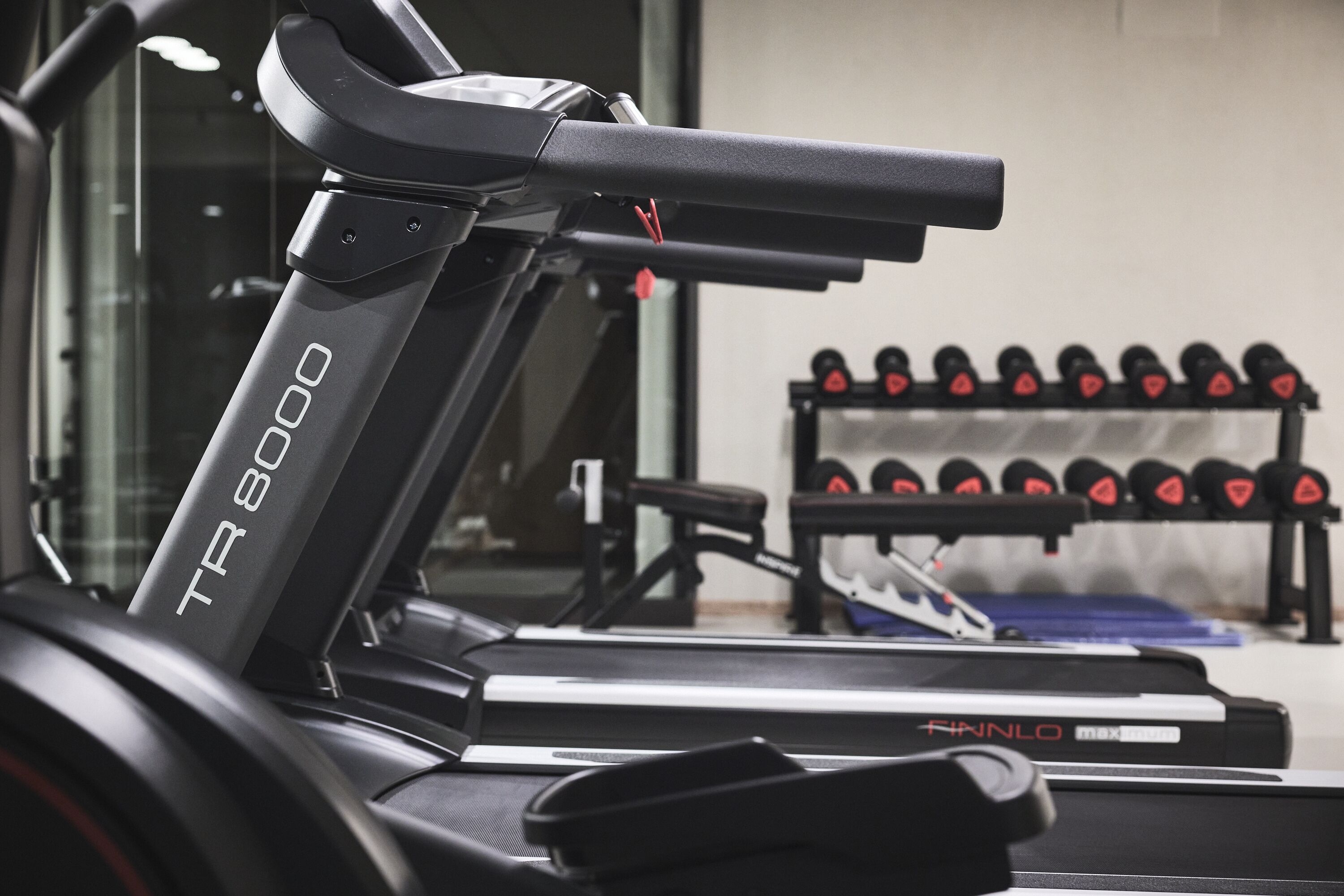 Un tapis de course TR 8000 avec un banc de musculation et des haltères en arrière-plan.