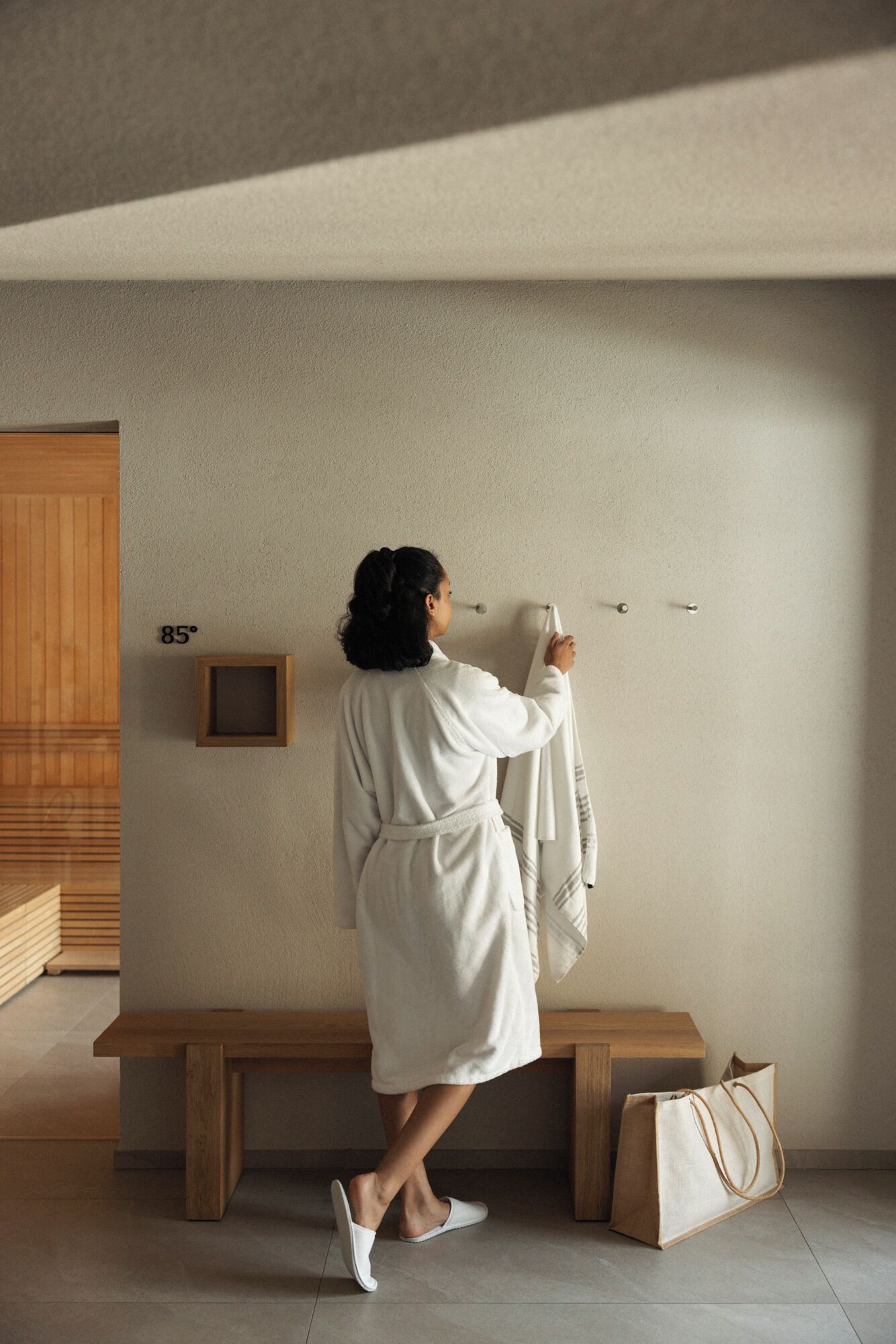 A person in a white robe hanging a towel on a wall-mounted towel rack in a room with a wooden bench and a small wooden box.