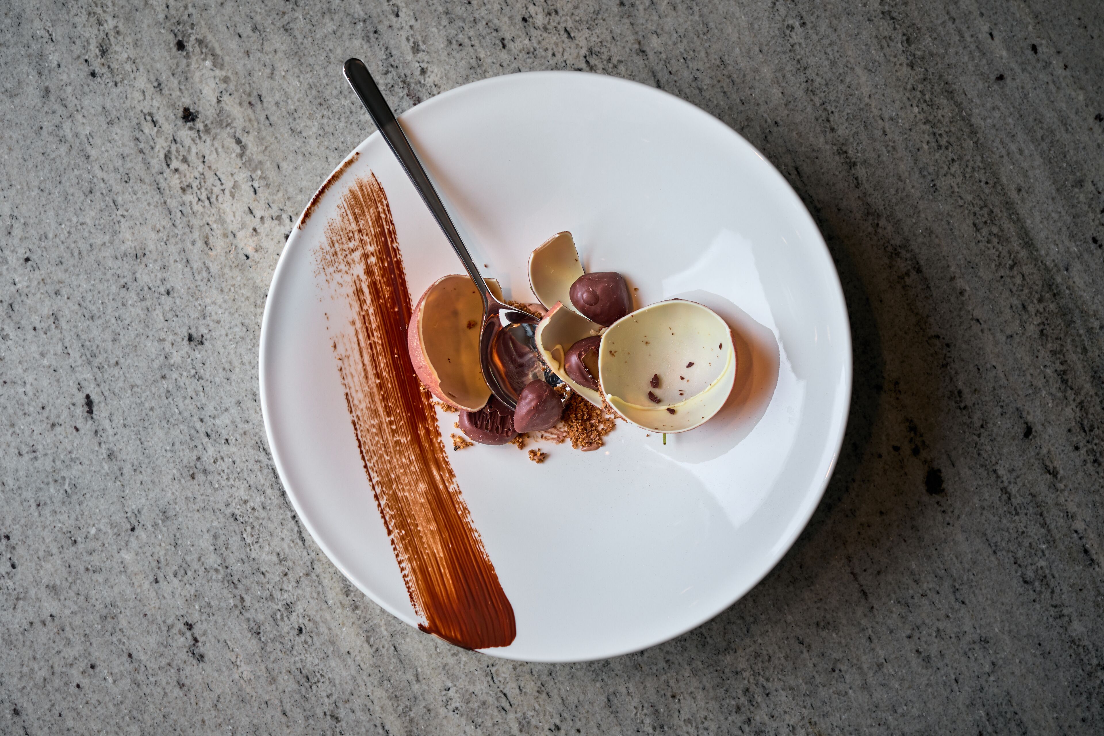 A white plate with a chocolate dessert, a spoon, and some fruit pieces on a speckled countertop.