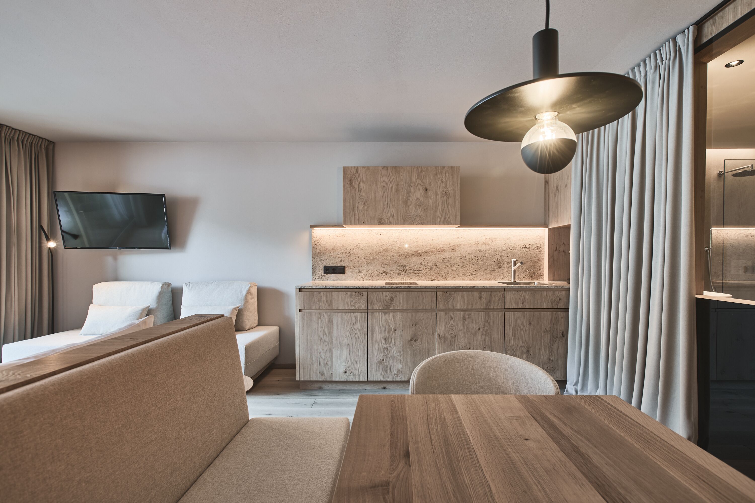A modern hotel room with a wooden dining table, a sofa, a TV mounted on the wall, and a kitchen area with a wooden countertop and cabinets.