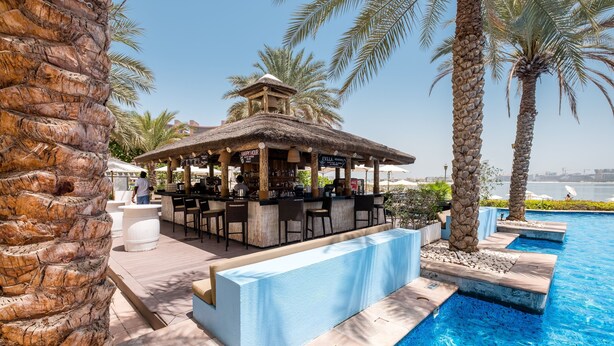 A poolside bar area with a thatched roof, seating, and a view of the pool. - Maison Privee - Stylish Apt with Large Patio close to the Beach (Dubai)