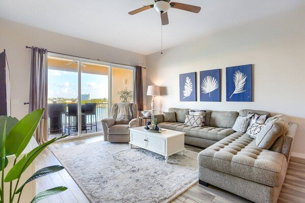 A living room with a sofa, a coffee table, a lamp, and a ceiling fan. There are three wall-mounted artworks and a sliding glass door leading to an outdoor area.
