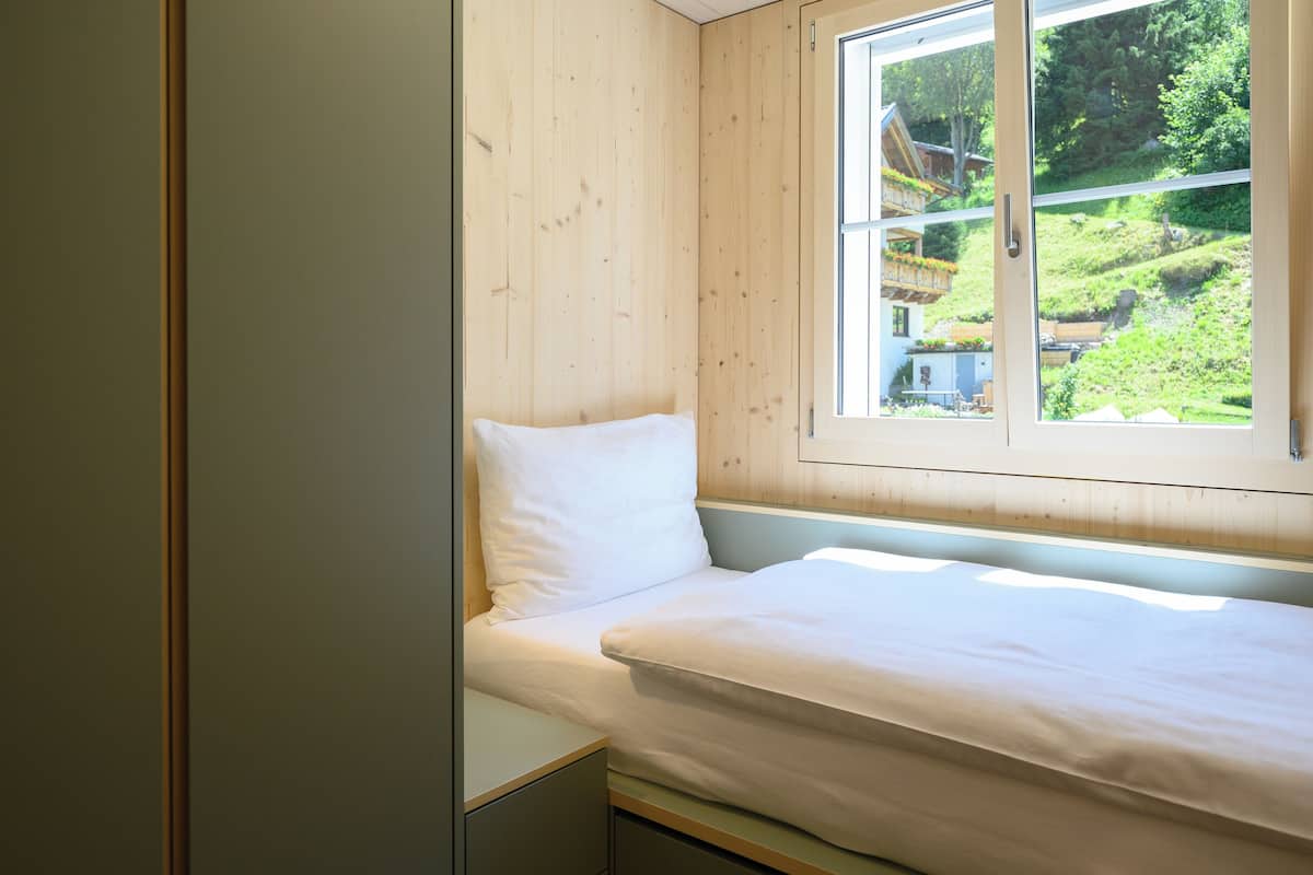 A bedroom with a wooden wall, a window with a view of a green landscape, and a bed with white bedding.