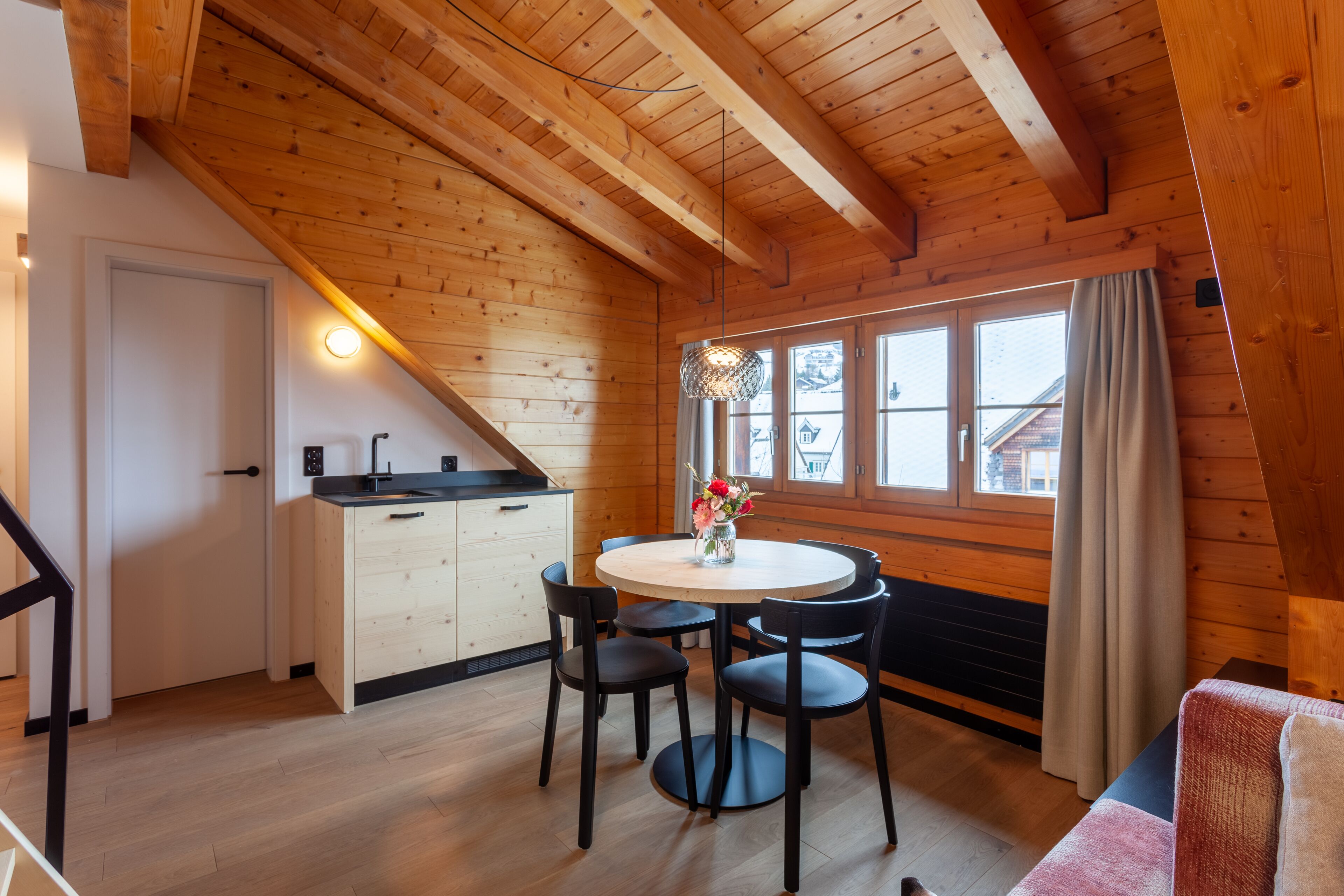 Une pièce chaleureuse avec un plafond en bois, une table de salle à manger ronde entourée de chaises, un petit coin cuisine et une fenêtre avec des rideaux.