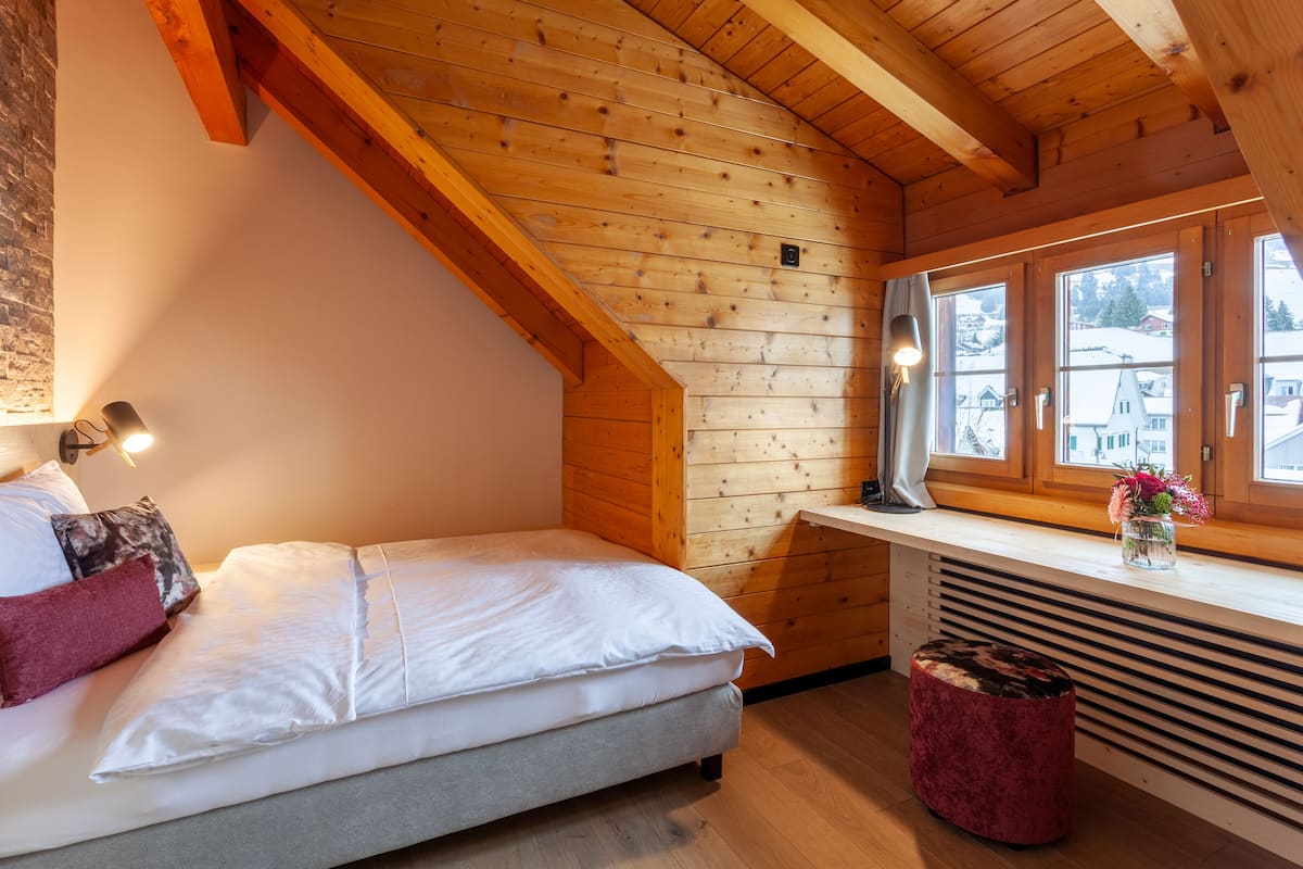 A cozy bedroom with a wooden ceiling, a bed with white bedding, a red ottoman, and a window with a view of buildings.