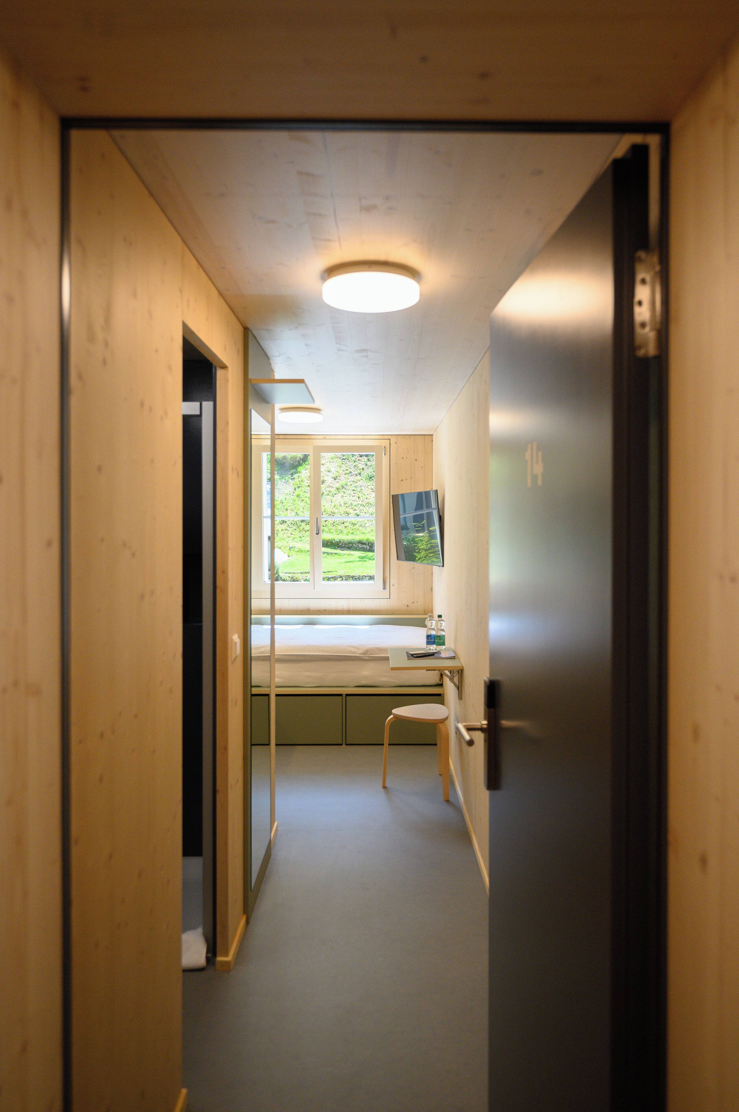 A narrow hallway with a wooden door on the right, a window with a view of greenery, and a small desk with a chair.