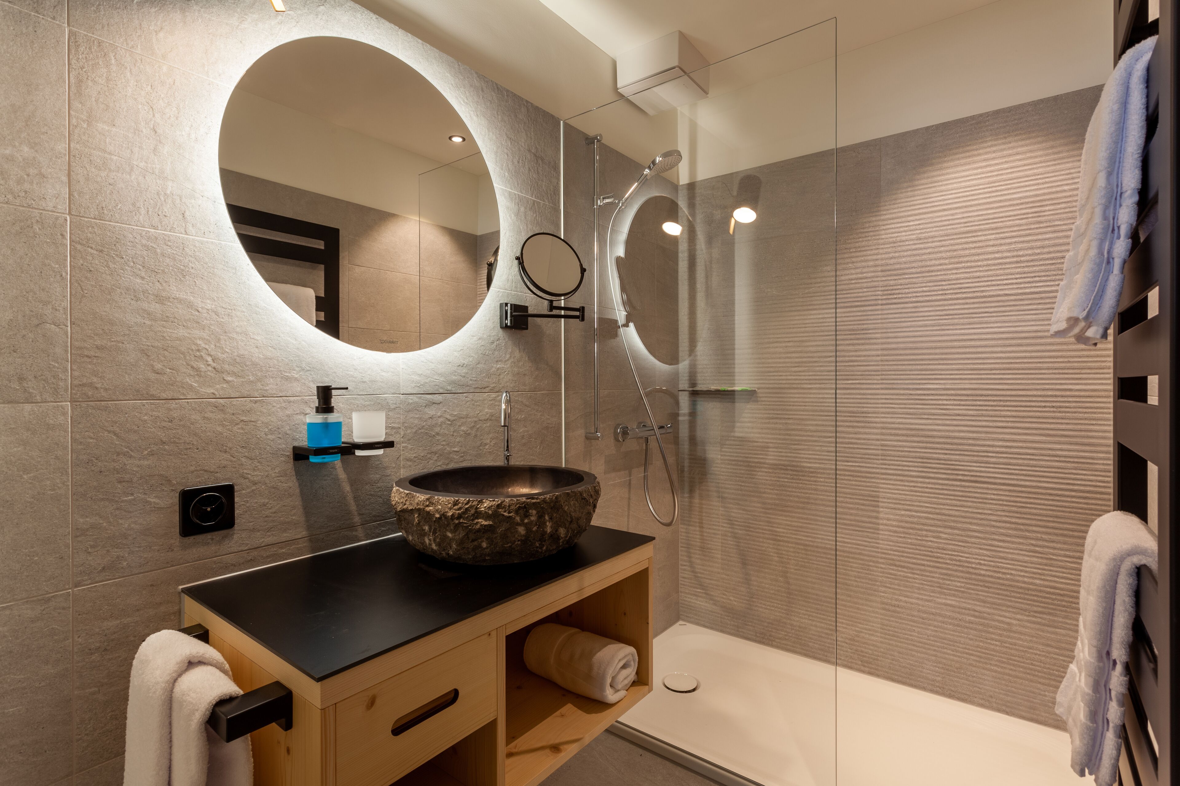 A modern bathroom with a large round mirror, a stone sink, a handheld showerhead, and a bathtub.
