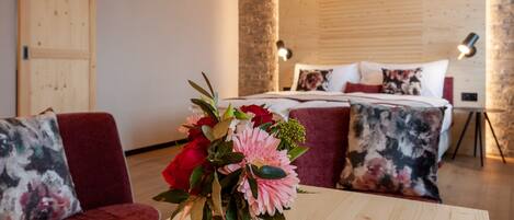A hotel room with a bed, a wooden headboard, a bedside table with a vase of flowers, and a red sofa with floral cushions.