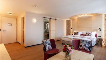 A modern hotel room with a wooden floor, a bed with white bedding, a red chair, a wooden table with a vase of flowers, and a sliding door leading to a bathroom.