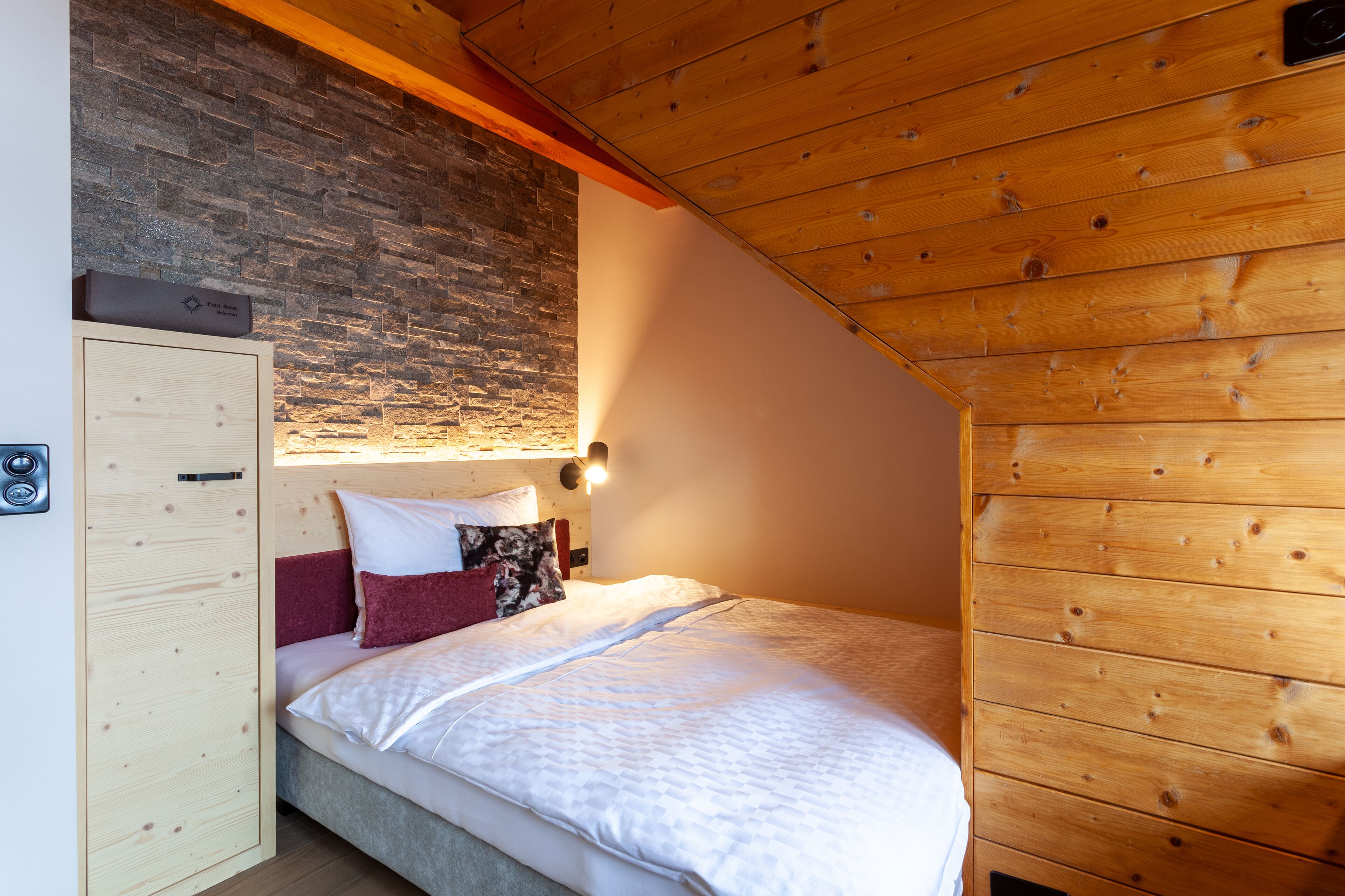 A bedroom with a sloped ceiling, wooden paneling, a stone wall, and a bed with white bedding and multiple pillows.