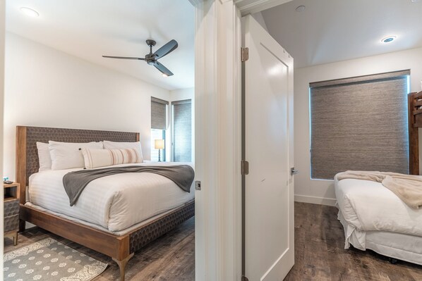 A bedroom with a bed, a nightstand, a ceiling fan, a window with blinds, and a door.