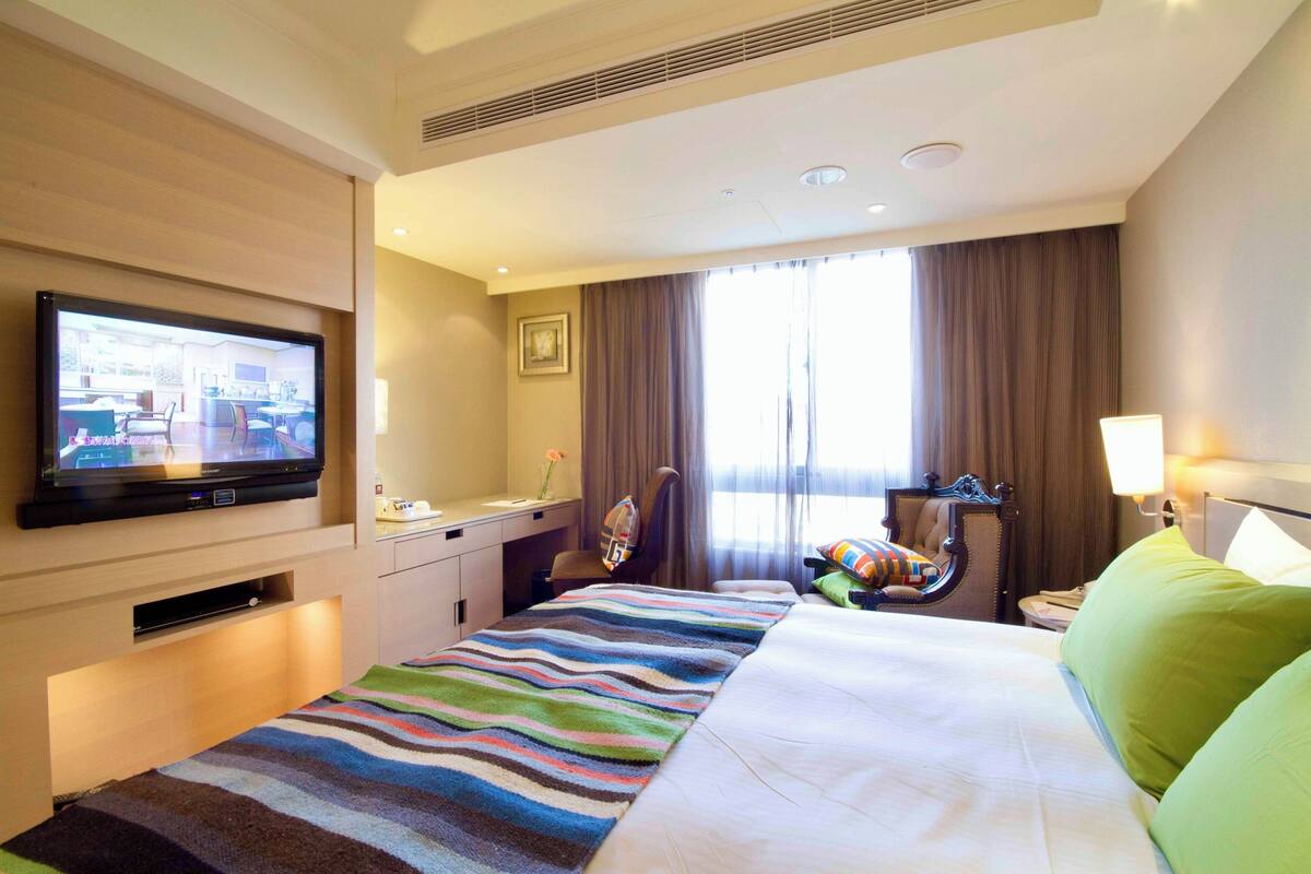 Ein Hotelzimmer mit einem großen Bett, einem Schreibtisch mit Stuhl, einem an der Wand befestigten Fernseher und einem Fenster mit Vorhängen.