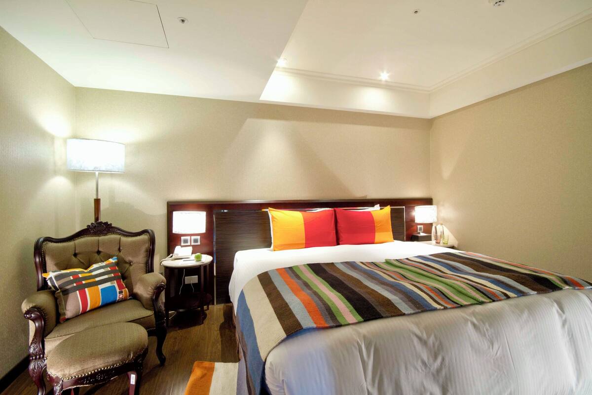 Ein Hotelzimmer mit einem großen Bett, einem Sessel mit bunten Kissen, einem kleinen Tisch und einer Lampe.