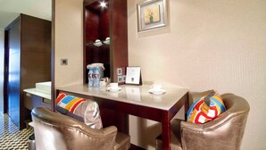 Een hotelkamer met een houten eettafel, twee leren stoelen, een klein wit tafeltje en een lijst met een afbeelding aan de muur.
