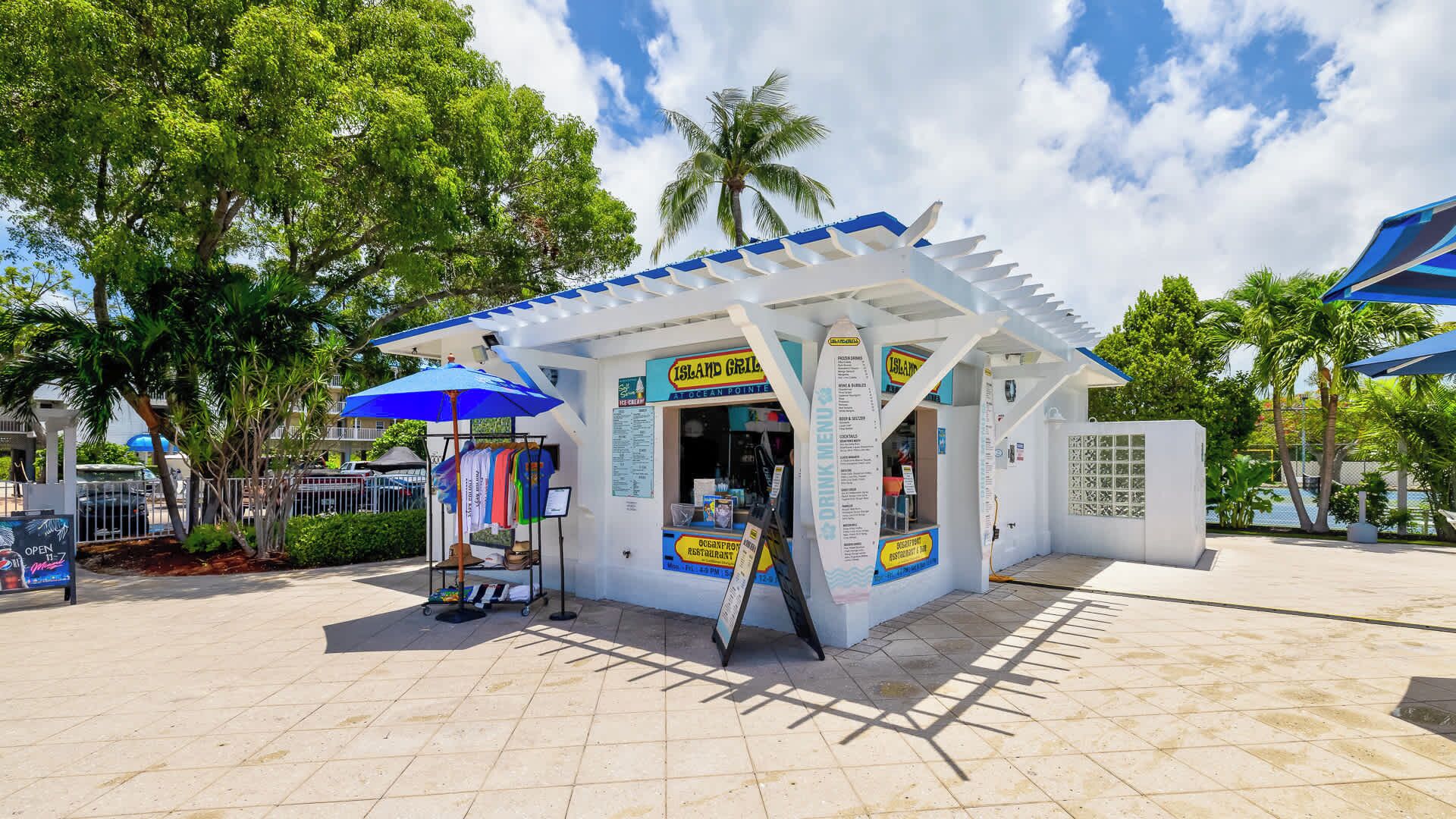 En lille, hvid kiosk med et blåt tag, med teksten 'Island Grill', der viser et menukort og et skilt til en restaurant ved stranden.