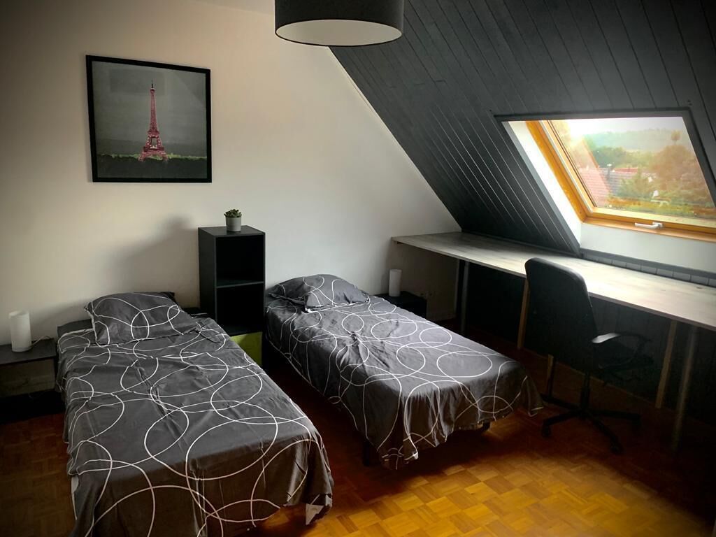 Une chambre avec deux lits, un bureau, une chaise, une fenêtre donnant sur un immeuble et une image de la Tour Eiffel accrochée au mur.