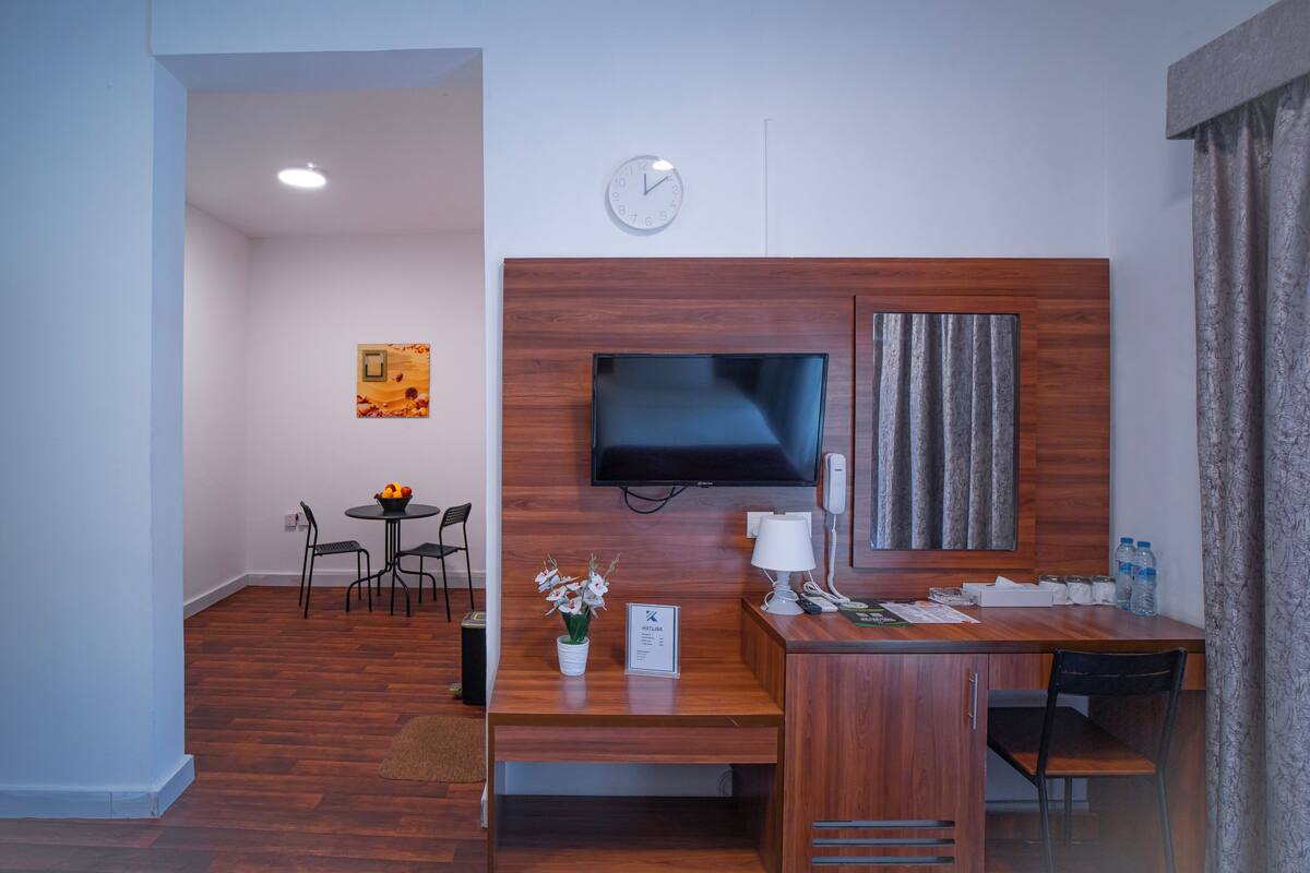 Een hotelkamer met een houten bureau, een flatscreen televisie aan de muur, een klok, een klein tafeltje met stoelen, en een schilderij.
