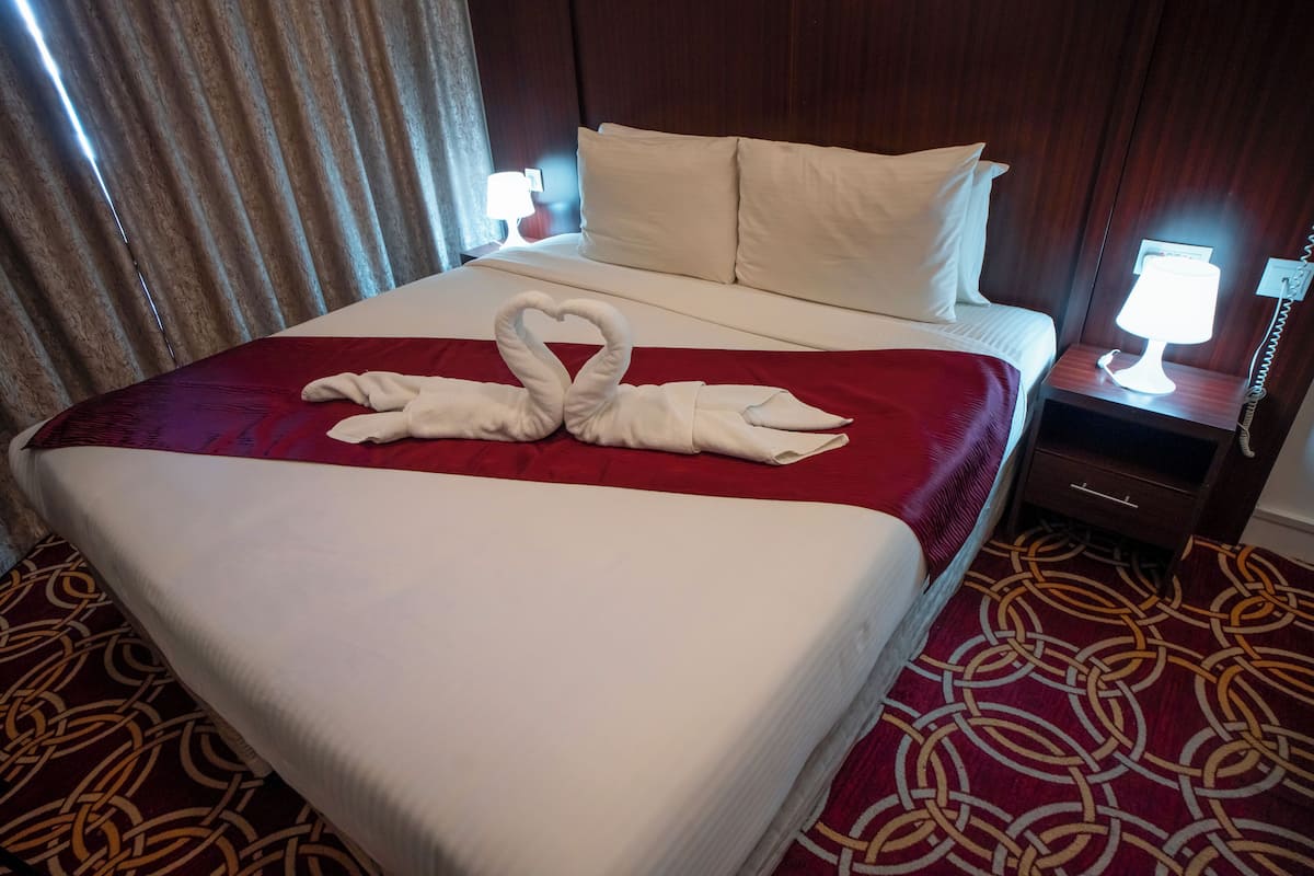 Een hotelkamer met een net opgemaakt bed, een handdoekarrangement in de vorm van een hart, een kamerbreed tapijt met een patroon, en een nachtkastje met een lamp.