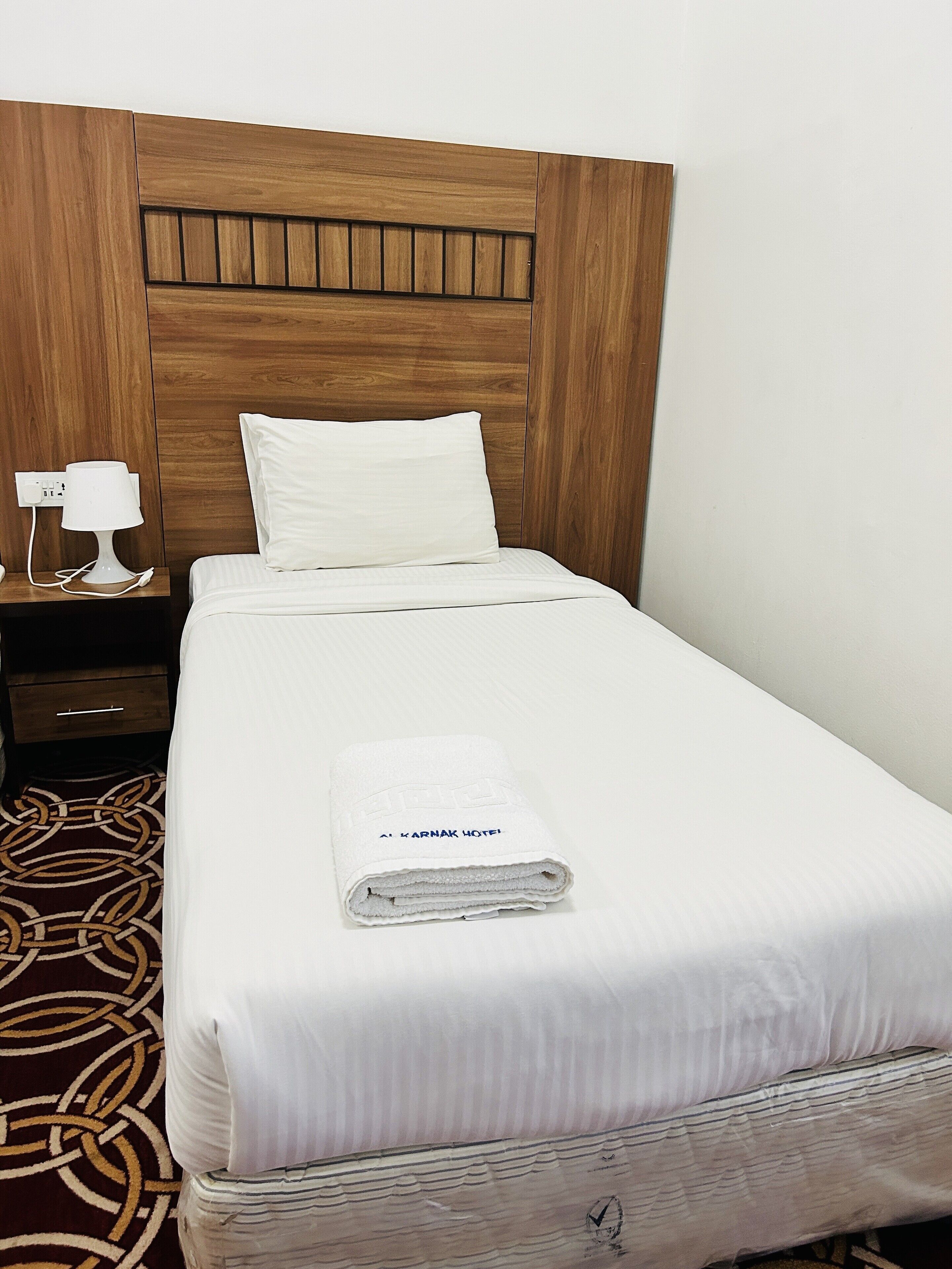 A neatly made bed with white linens, a folded towel with text, a bedside lamp, and a wooden headboard.