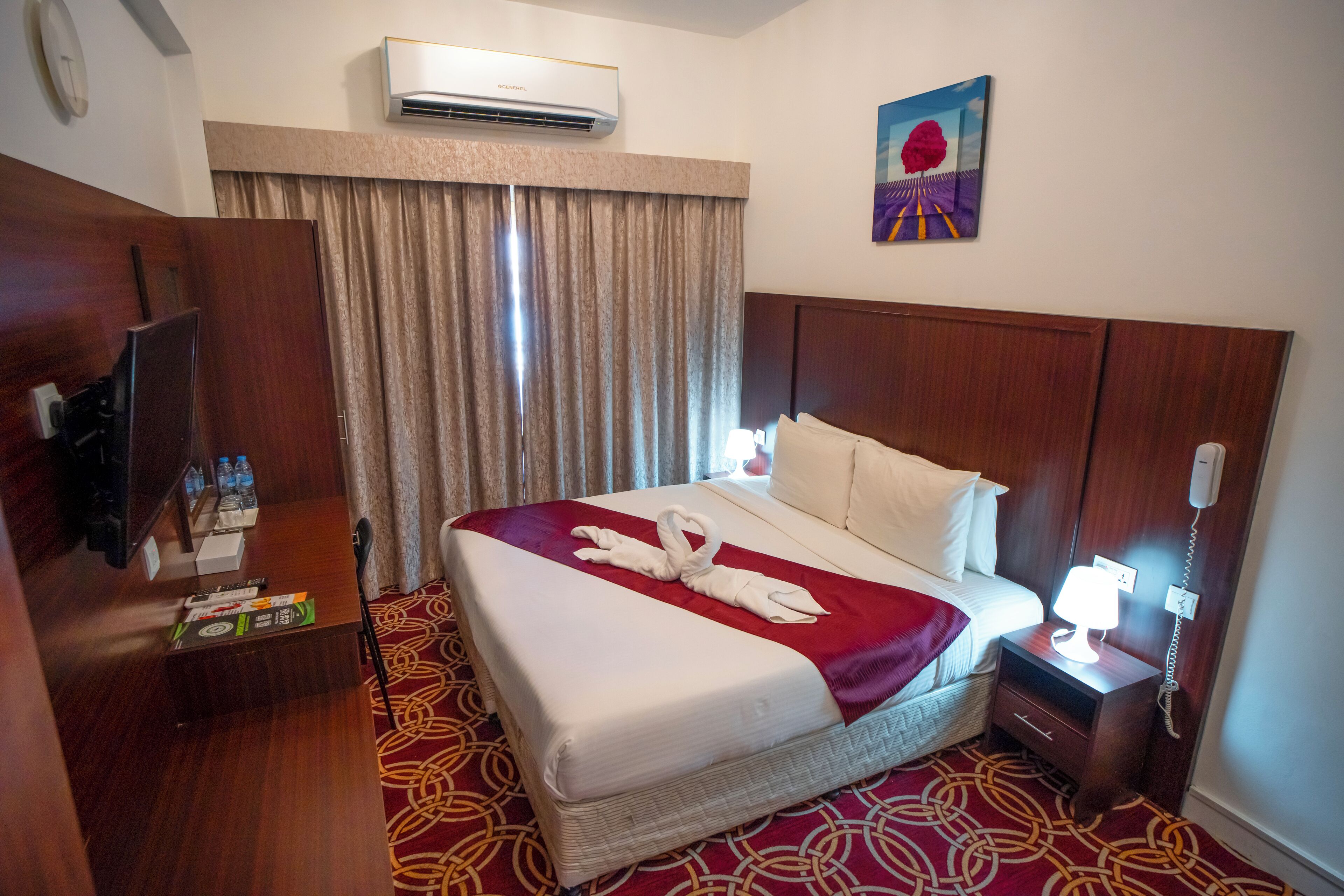 A hotel room with a bed, a TV, a minibar, a desk, a telephone, and an air conditioning unit.