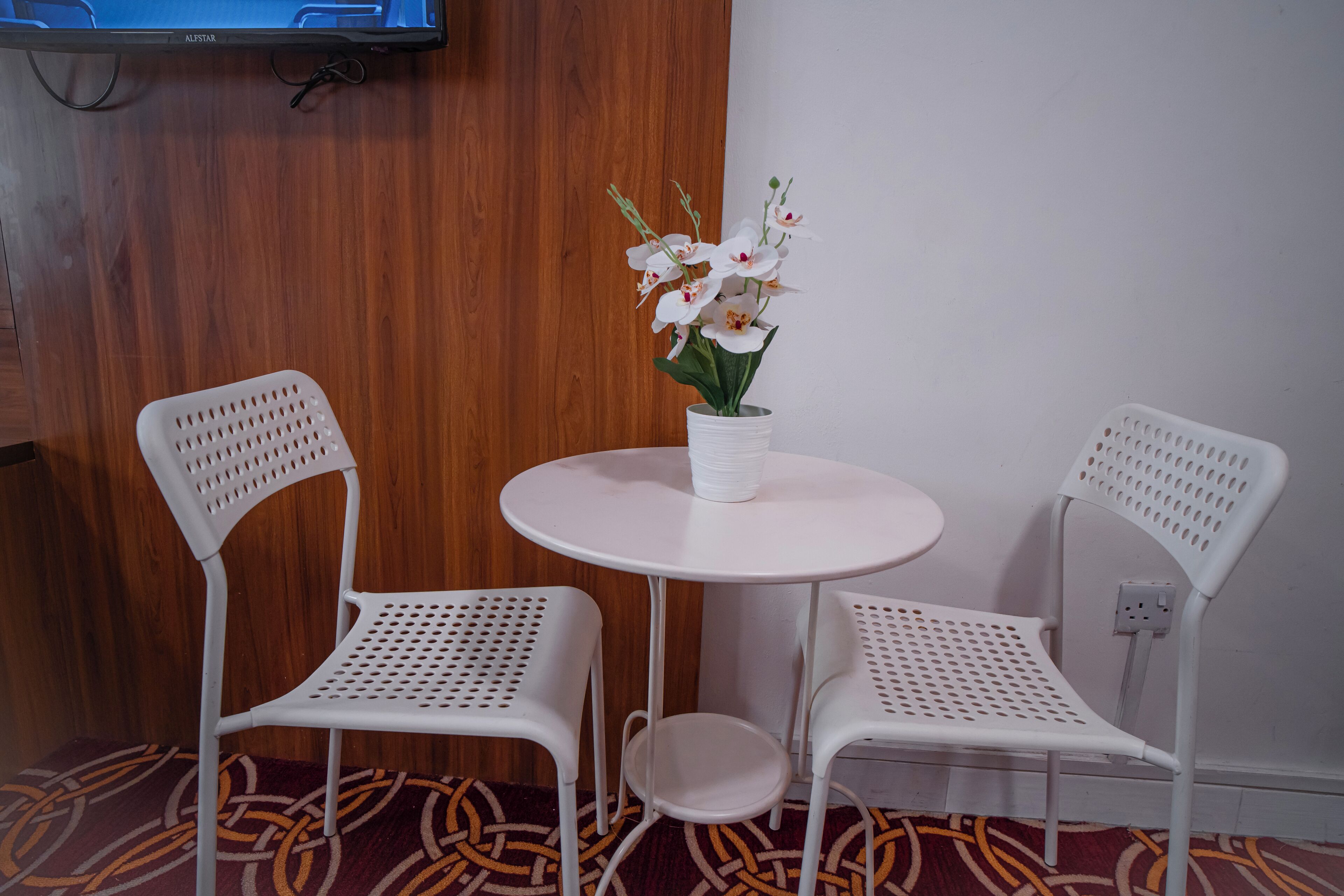 Une petite table blanche, accompagnée de deux chaises blanches, un vase blanc contenant des fleurs blanches, et un mur en bois sur lequel est fixé un téléviseur.