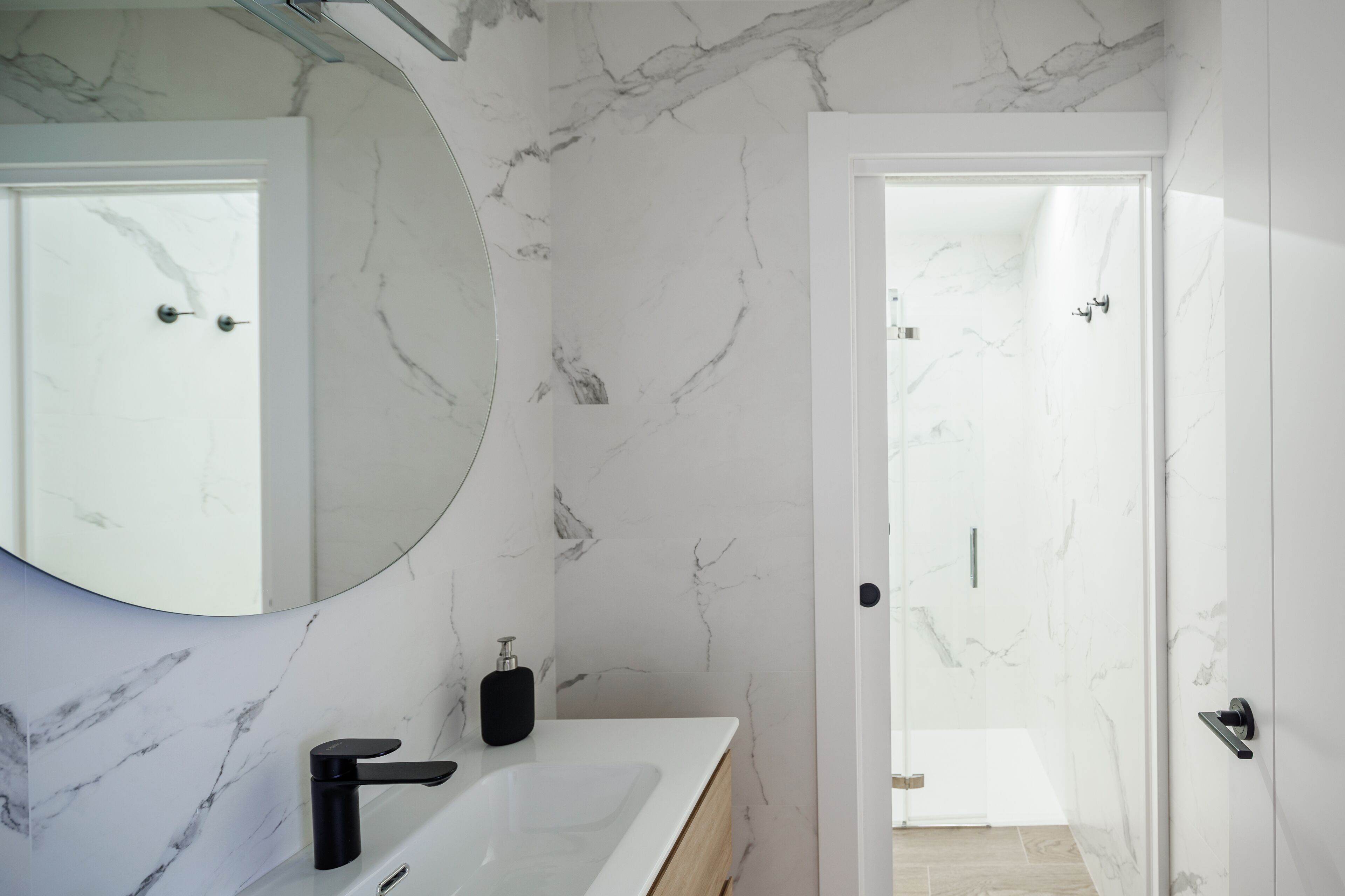 Ein modernes Badezimmer mit Marmorwand, einem großen Spiegel, einem weißen Waschbecken und einem schwarzen Wasserhahn.