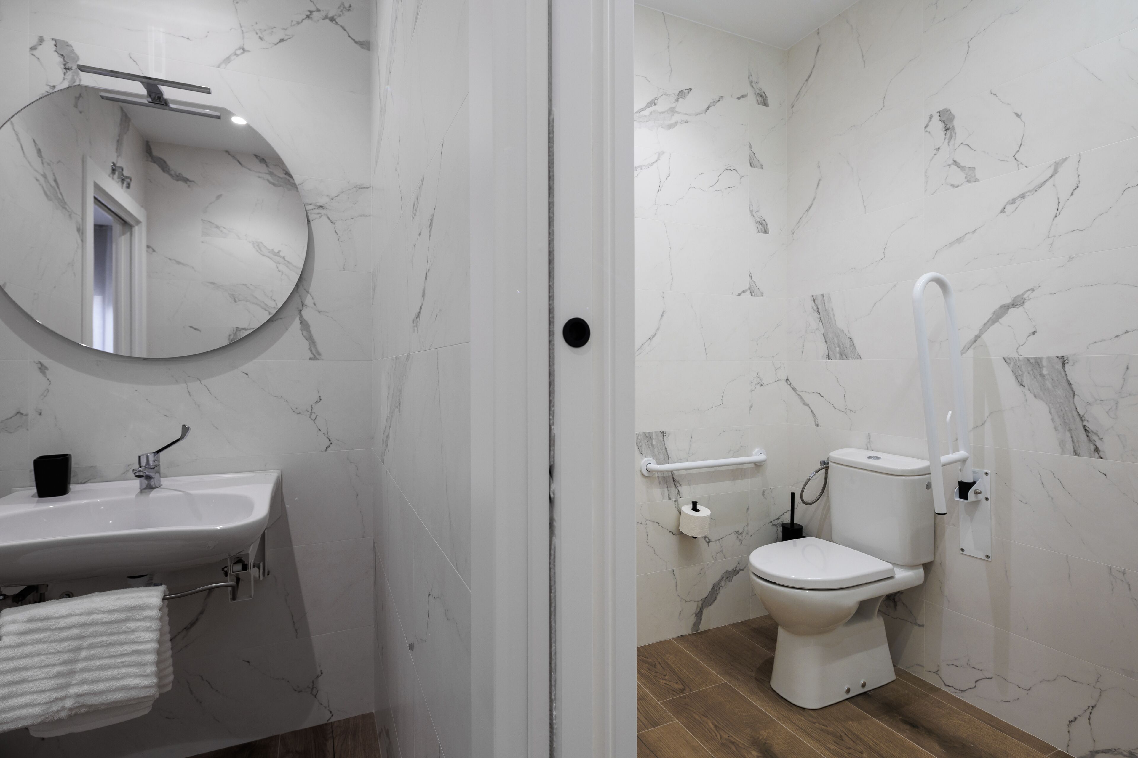 Un baño con pared de azulejos de mármol, un lavabo blanco con una toalla y un inodoro con barra de apoyo.