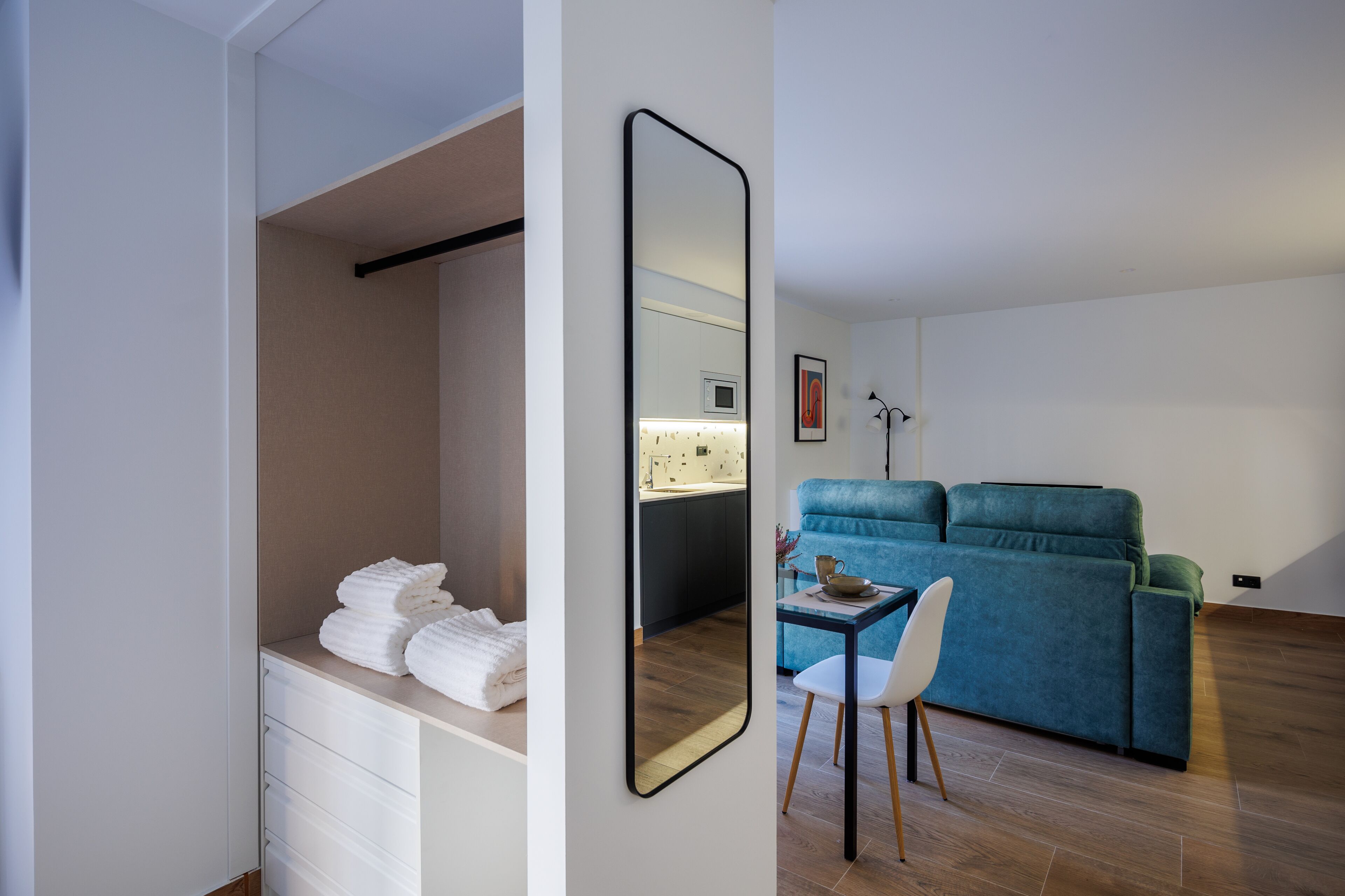 A modern hotel room with a large mirror, a kitchenette, a blue sofa, and a dining area with a chair.