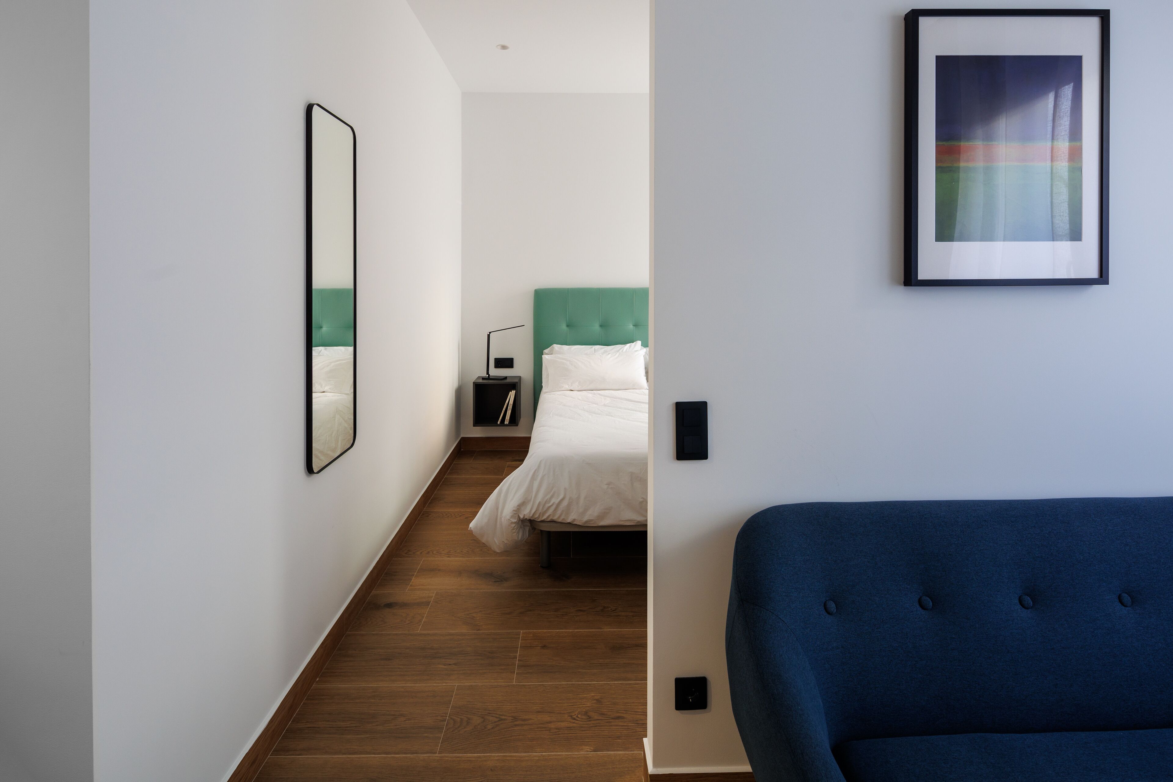 A modern hotel room with a wooden floor, a green headboard, a white bed, a blue sofa, a framed abstract painting, and a mirror on the wall.