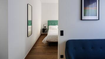 A modern hotel room with a wooden floor, a green headboard, a white bed, a blue sofa, a framed abstract painting, and a mirror on the wall.